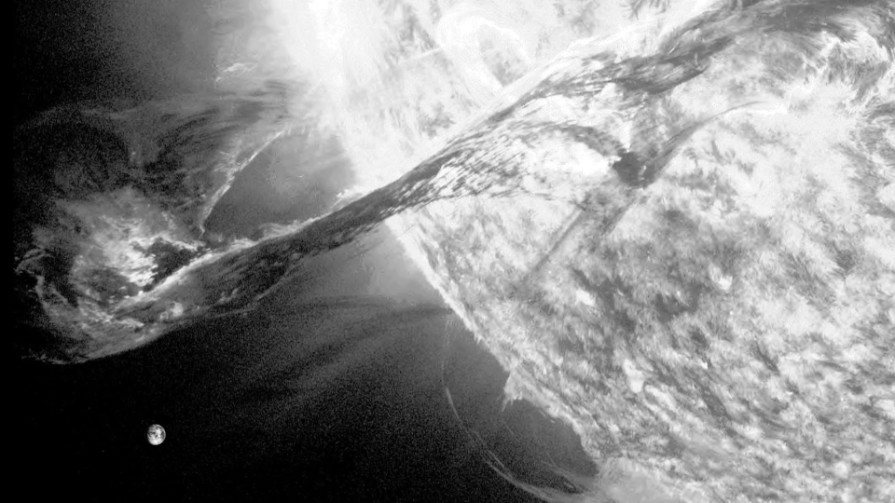 Black-and-white still: a glaring arc of light above a grainy, curved surface — a prominence at the edge of the sun.