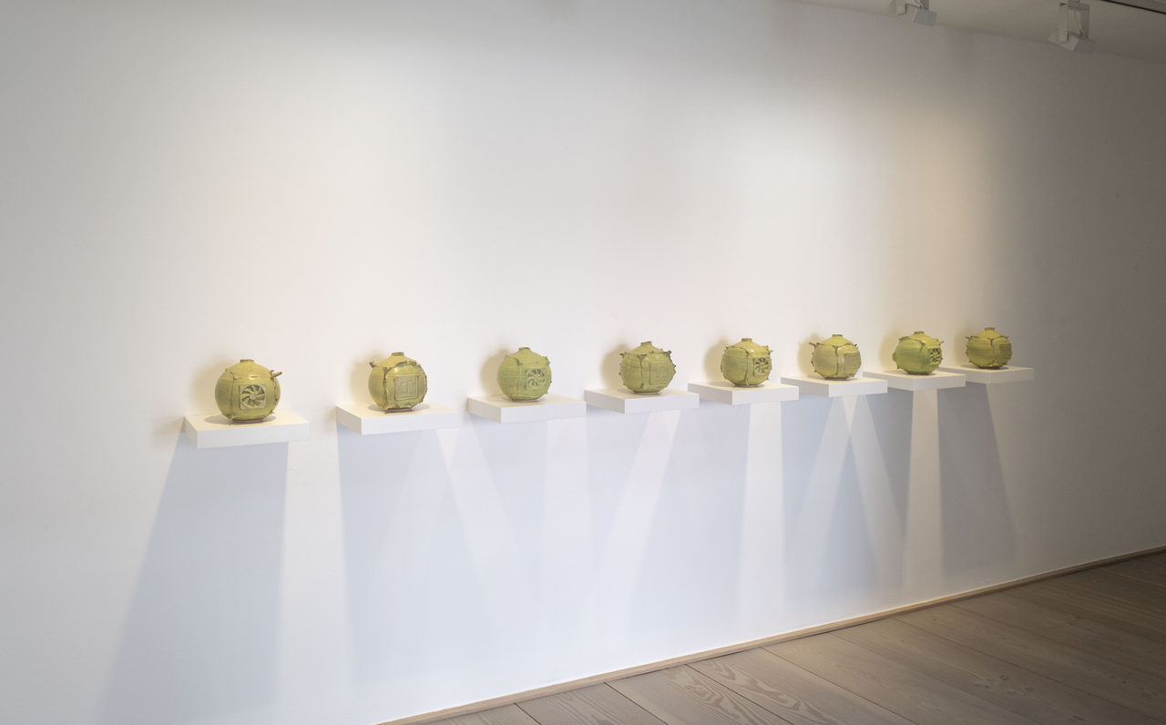 Eight bellied, yellow-green glazed ceramic vessels stand one to a white wall-mounted plinth in a slightly rising row, casting long shadows on the wall.