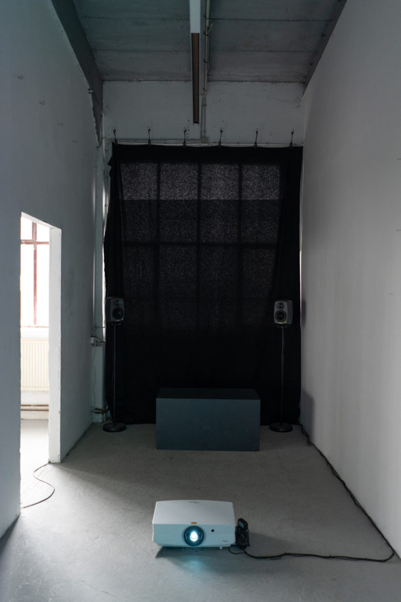Installation view: a dark curtain serving as projection surface spans the end wall of a room, with two floor speakers, a low blue plinth and a projector standing on the floor in front of it.
