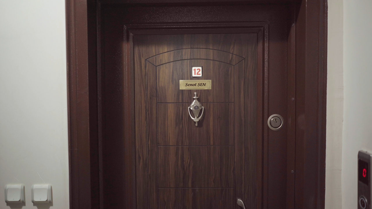 Still: a dark wooden apartment door with a house number, a nameplate and a bell, two white meter boxes beside it.