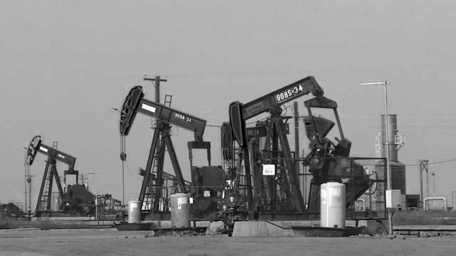Black-and-white still: a field of oil pumpjacks and storage tanks under an open sky.