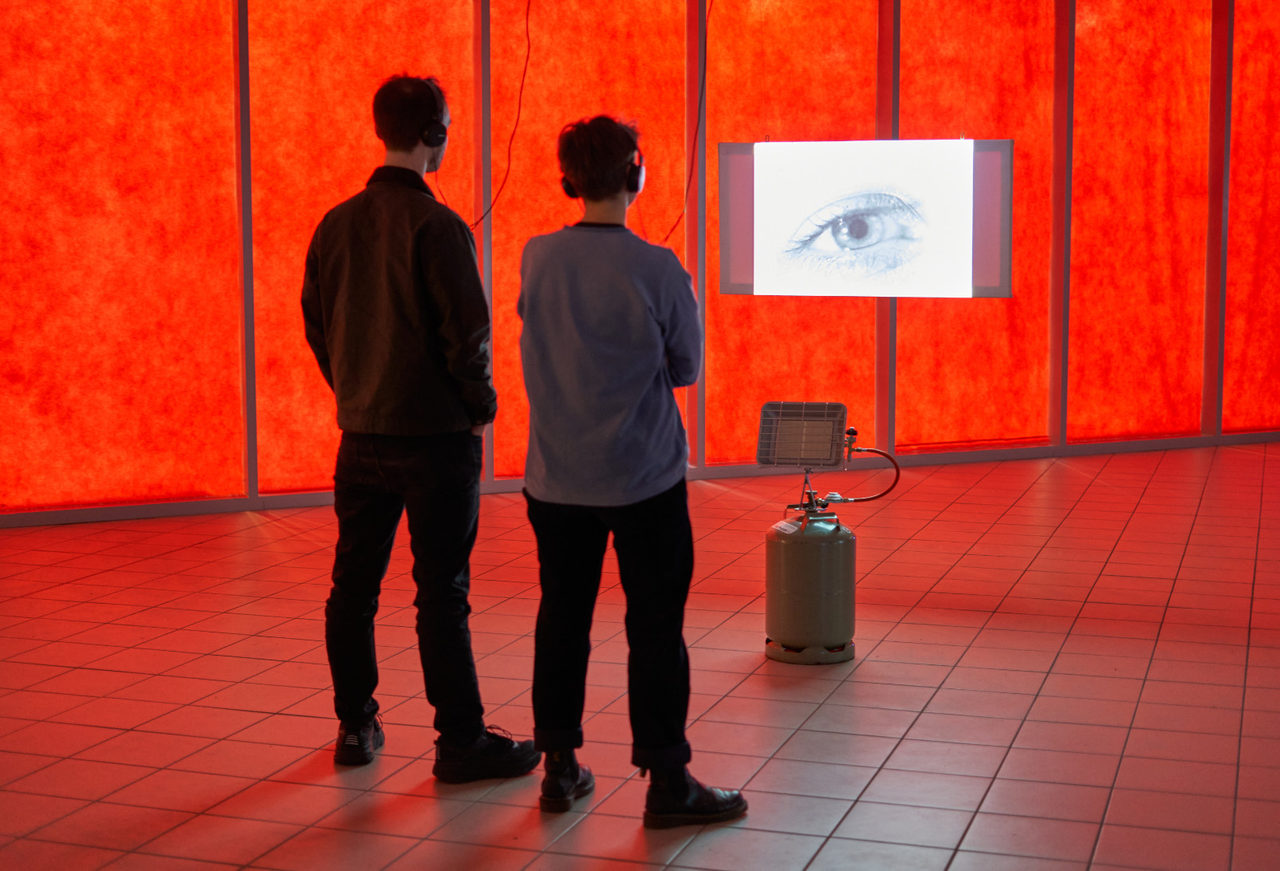 Installation view: two people stand with their backs to the camera in a red-lit room in front of a small suspended screen showing an eye; a gas patio heater stands beside it.