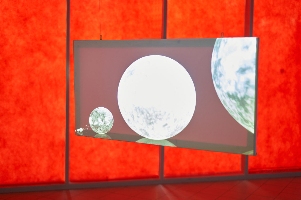 Detail: a semi-transparent screen suspended on two wires in front of a red glowing wall, showing bright spheres of different sizes against a dark ground.