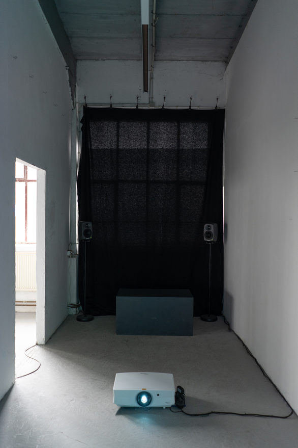 Installation view: a dark curtain serving as projection surface spans the end wall of a room, with two floor speakers, a low blue plinth and a projector standing on the floor in front of it.