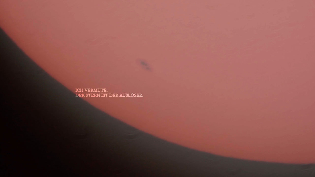 Still: a red-glowing solar disc with a dark sunspot fills most of the frame, with darkness at the lower left and two small lines of text.