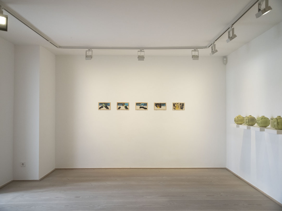 A bright gallery room. Five small framed ceramic panels hang in a row on the end wall; along the right-hand wall a line of yellow-green ceramic vessels stands on shelf plinths.