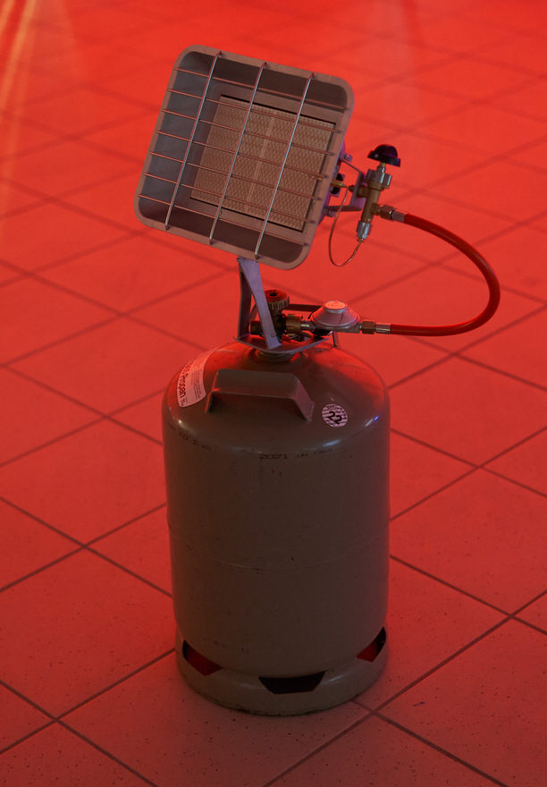 Detail: a gas radiant heater with its bottle and red hose on a red tiled floor in front of a red glowing wall.