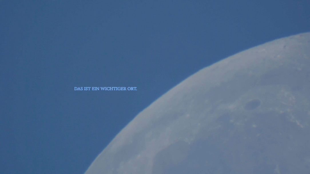 Still: the sunlit edge of a moon against a pale blue ground, with a small line of overlaid text above it.