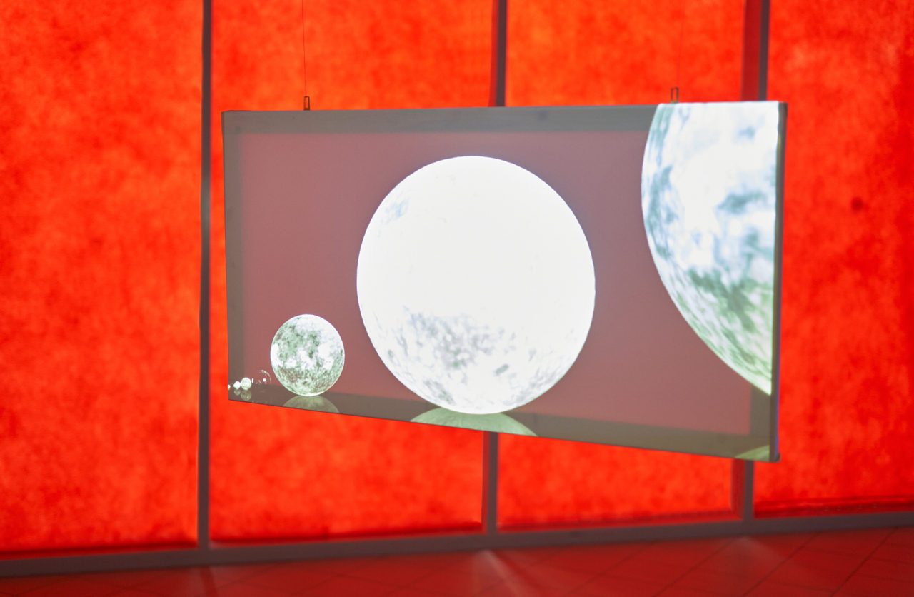 Detail: a semi-transparent screen suspended on two wires in front of a red glowing wall, showing bright spheres of different sizes against a dark ground.