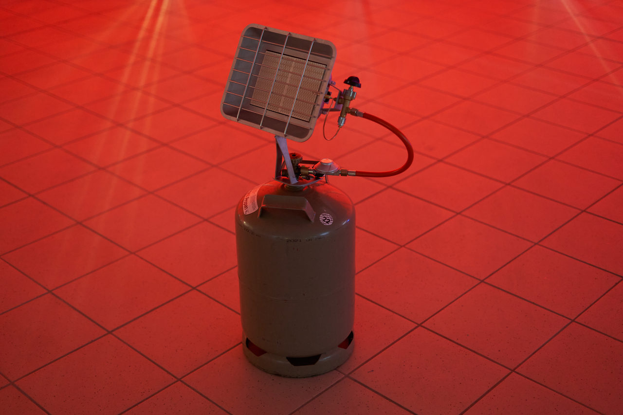 Detail: a gas radiant heater with its bottle and red hose on a red tiled floor in front of a red glowing wall.