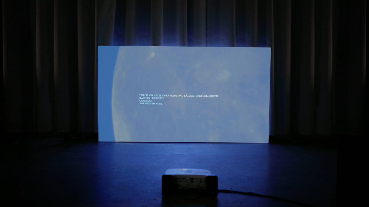 Installation view in a darkened hall: a large projection on a free-standing screen in front of a stage curtain, a projector on the floor before it; the projection shows a moon with text overlaid.
