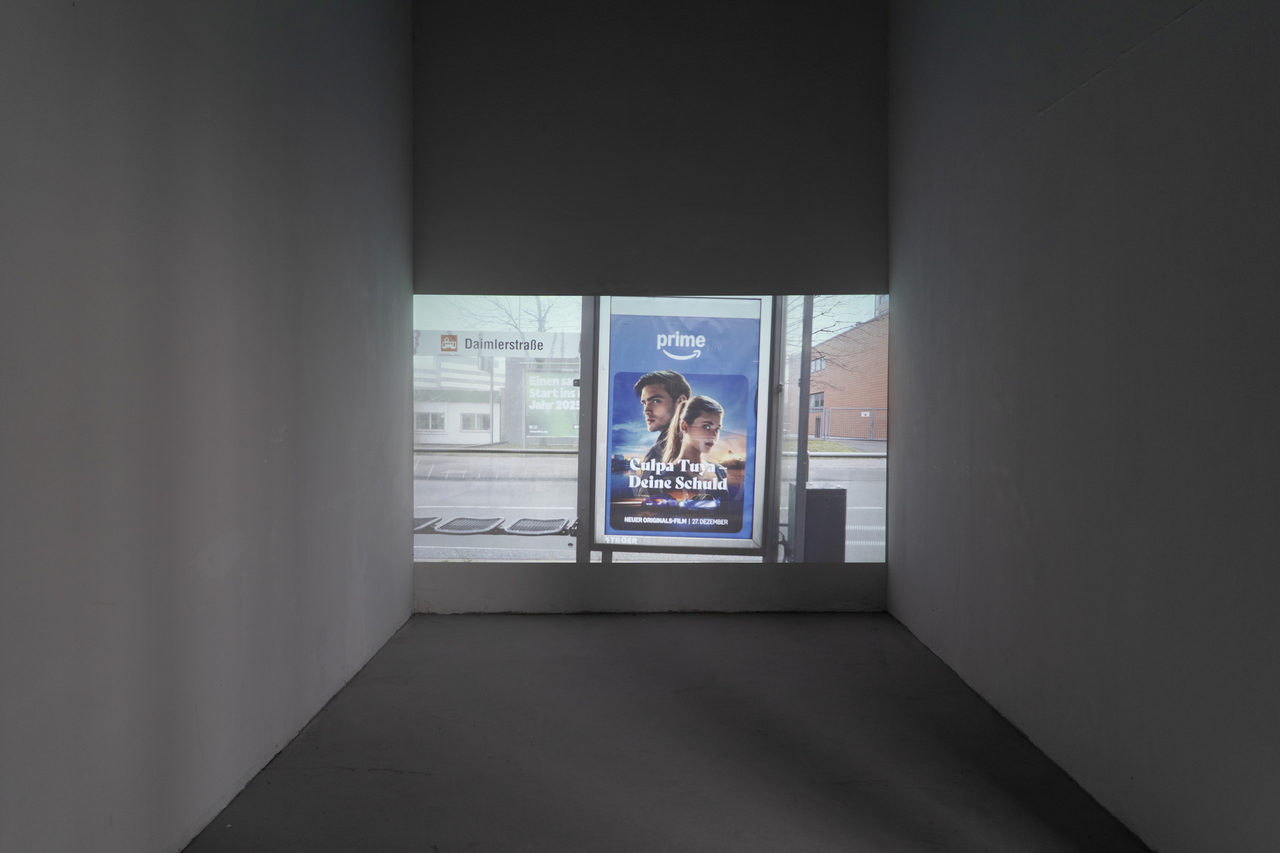 A darkened booth with a video projected onto its back wall: the view out of a window onto a street with a bus stop, parked cars and a lit advertising display.
