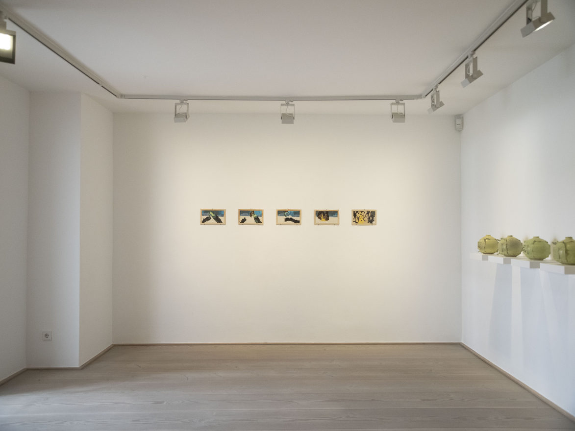 A bright gallery room. Five small framed ceramic panels hang in a row on the end wall; along the right-hand wall a line of yellow-green ceramic vessels stands on shelf plinths.