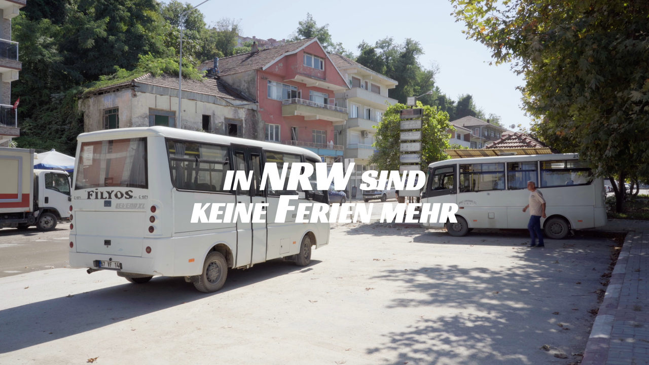 Still with the film title overlaid: two white minibuses on a dusty square, with a derelict stone house and a pink apartment building behind them.