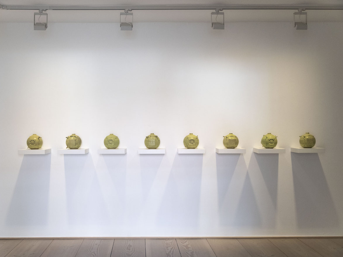 Eight bellied, yellow-green glazed ceramic vessels stand one to a white wall-mounted plinth in an even row, casting long shadows on the wall.