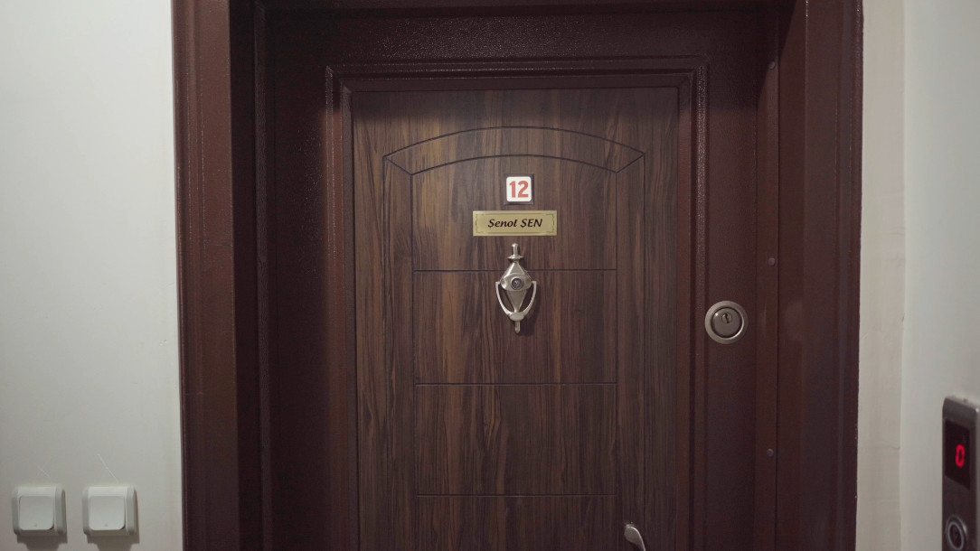 Still: a dark wooden apartment door with a house number, a nameplate and a bell, two white meter boxes beside it.