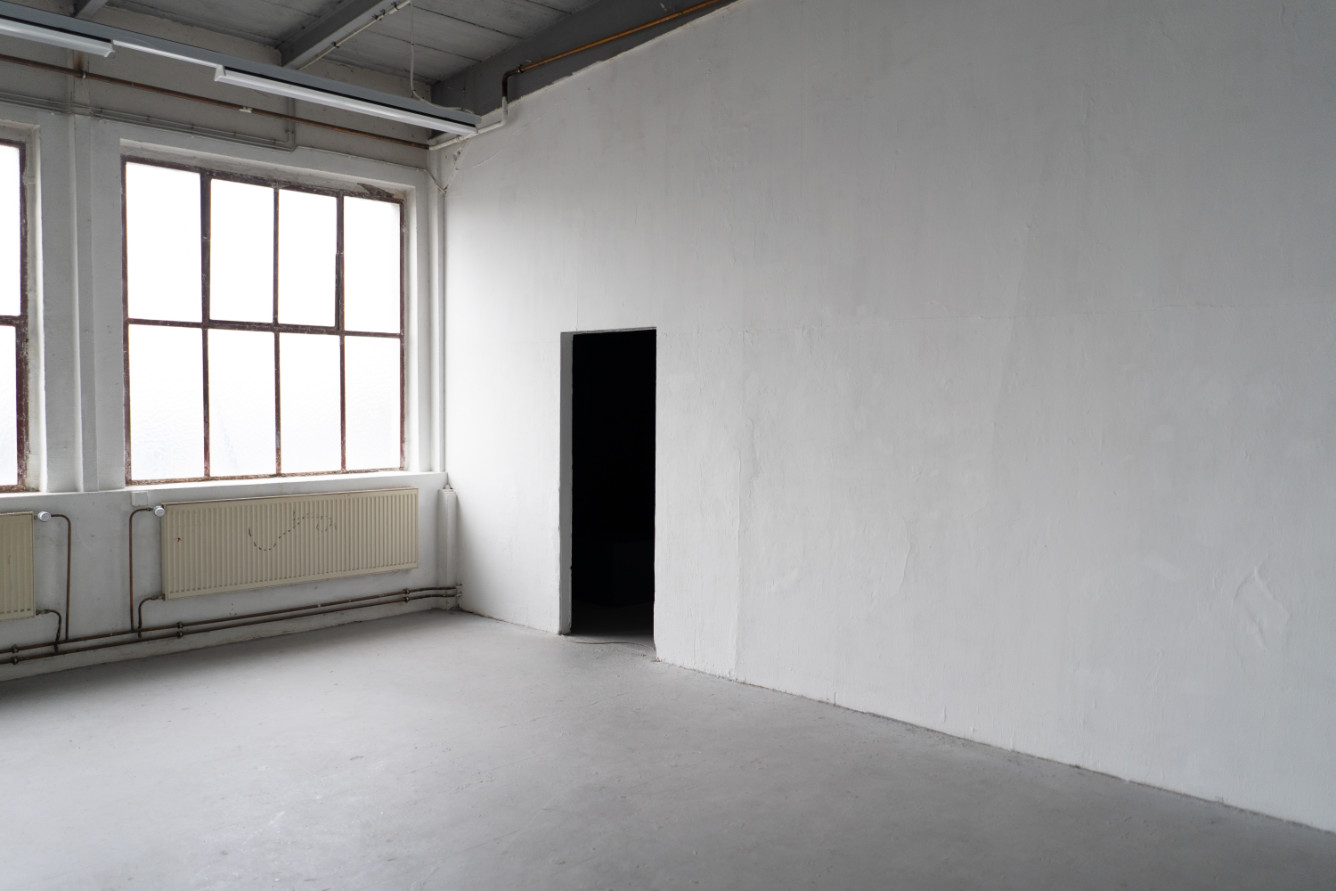 An empty studio room with tall multi-pane windows, a grey screed floor and an open, dark doorway in the whitewashed wall.